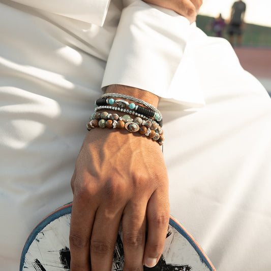 A close-up of a person wearing a white thobe and multiple beaded bracelets on one wrist, with a skateboard resting on their lap in an outdoor setting.