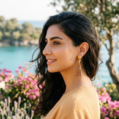 Woman with dark hair and earrings standing outdoors with flowers and water in the background